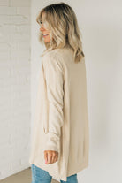 A woman wearing a long cardigan with pockets, paired with a white top and blue jeans, standing in front of a white brick wall.