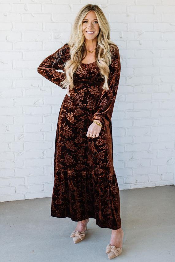 Woman wearing a brown patterned dress against a white brick wall.