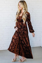 Woman wearing a brown velvet dress against a white brick wall.