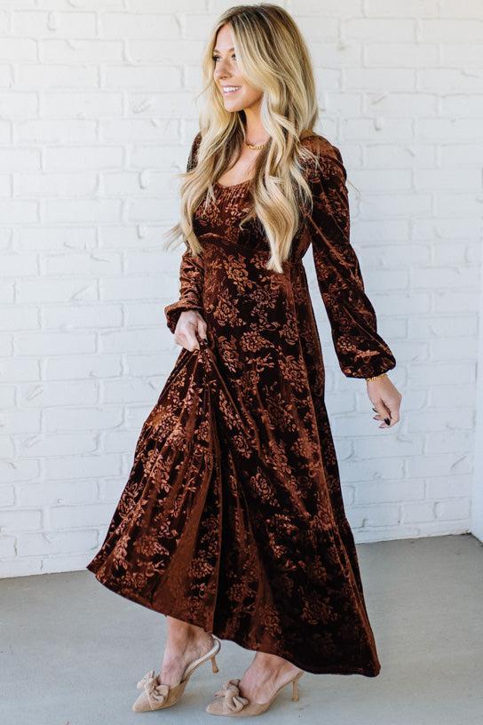 Woman wearing a brown velvet dress against a white brick wall.