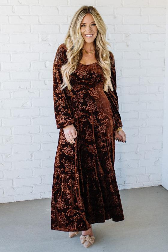 Woman wearing a brown floral dress against a white brick wall.