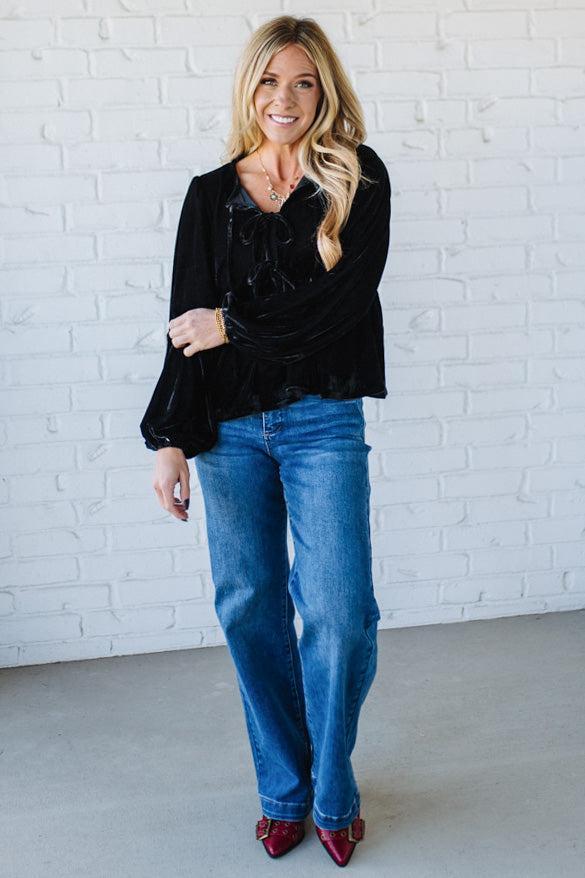 Woman wearing a black blouse and blue jeans against a white brick wall.