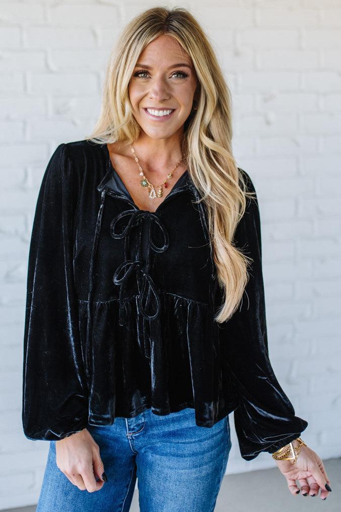Woman wearing a black velvet top and blue jeans against a white brick wall.
