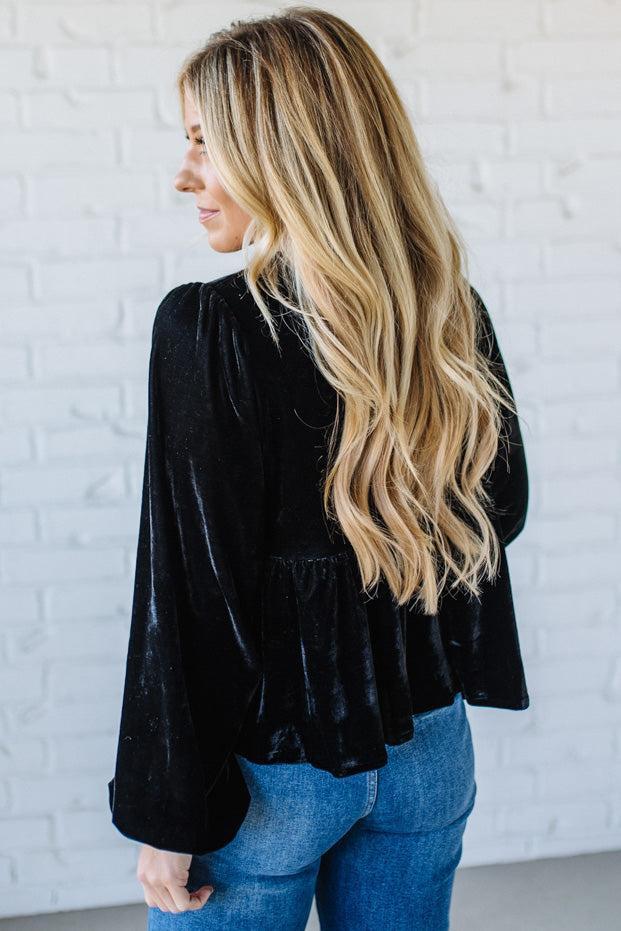 Woman wearing a black velvet top and blue jeans against a white brick wall.