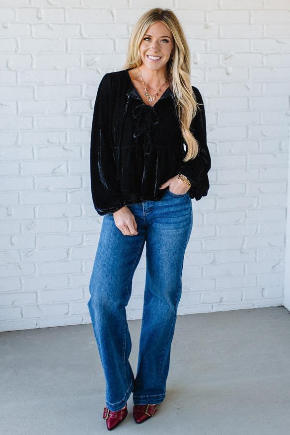 Woman wearing a black top and blue jeans against a white brick wall.