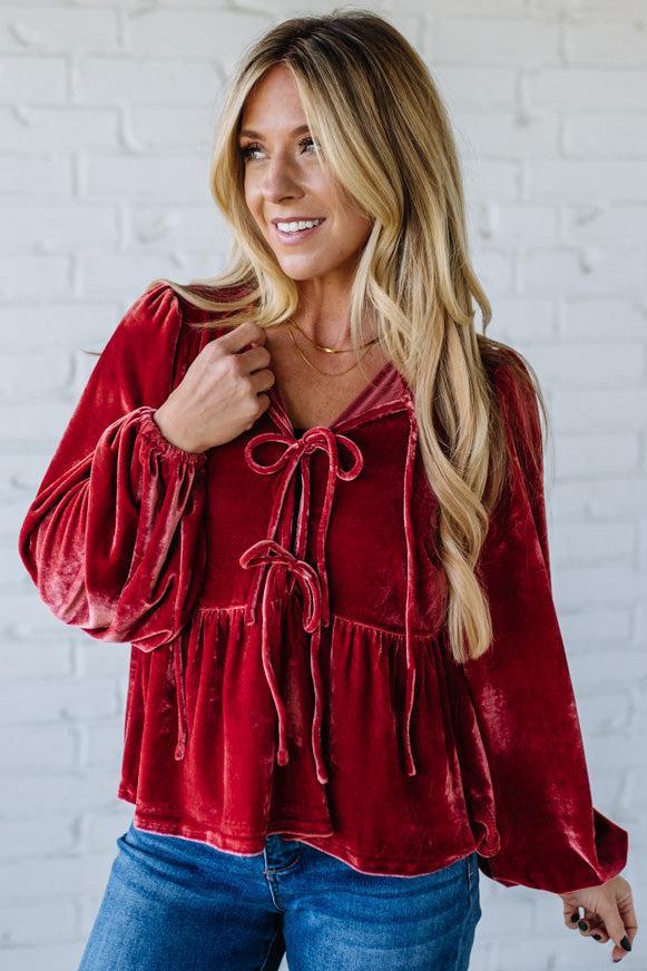 Woman wearing a red velvet top with blue jeans against a white brick wall.