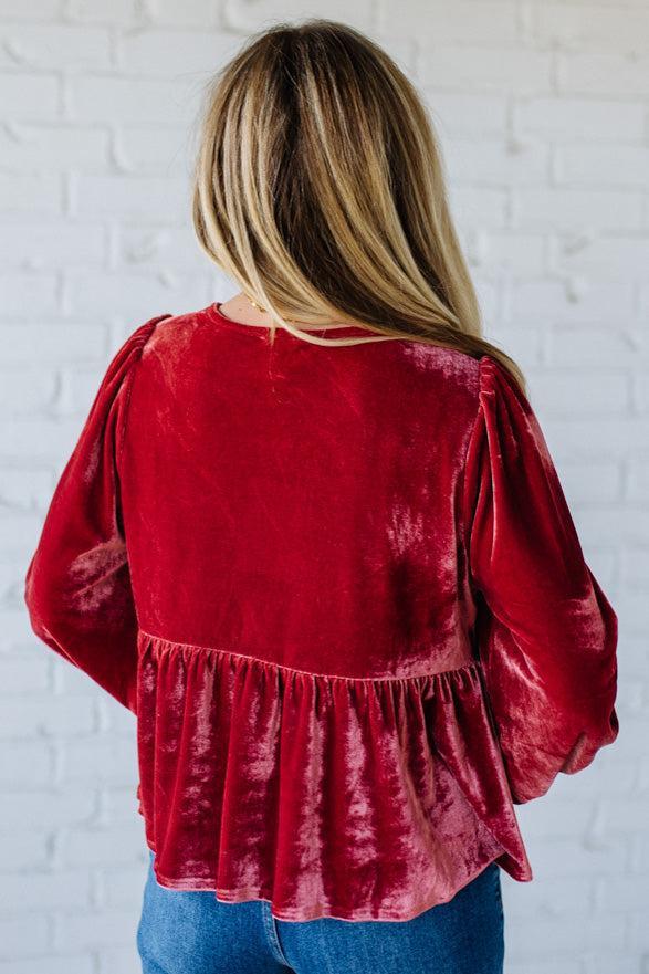 Red velvet blouse with long sleeves worn by a person against a white brick wall.