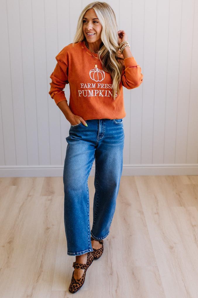 Blonde woman wearing a rust orange colored corduroy sweatshirt with white embroidered pumpkin and text: FARM FRESH PUMPKINS