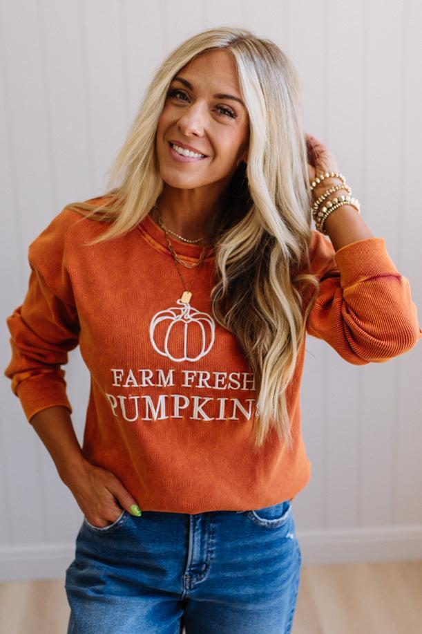 Blonde woman wearing a rust orange colored corduroy sweatshirt with white embroidered pumpkin and text: FARM FRESH PUMPKINS
