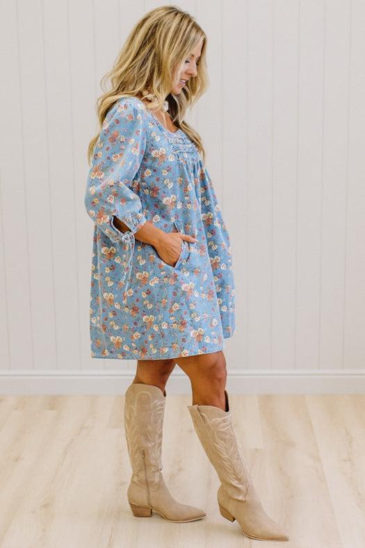 A woman wearing a light blue floral long-sleeve dress with pockets, paired with beige cowboy boots, standing in a room with white paneled walls and light wood floors.