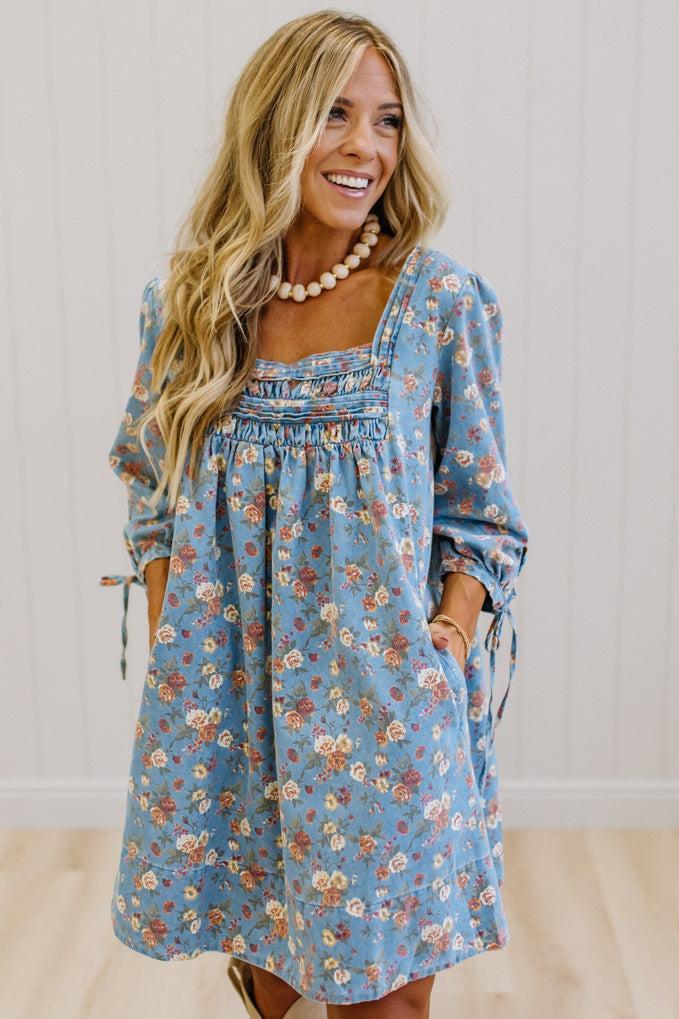 A woman wearing a light blue floral long-sleeve dress with a smocked bodice and tie details on the sleeves, standing against a white paneled wall.