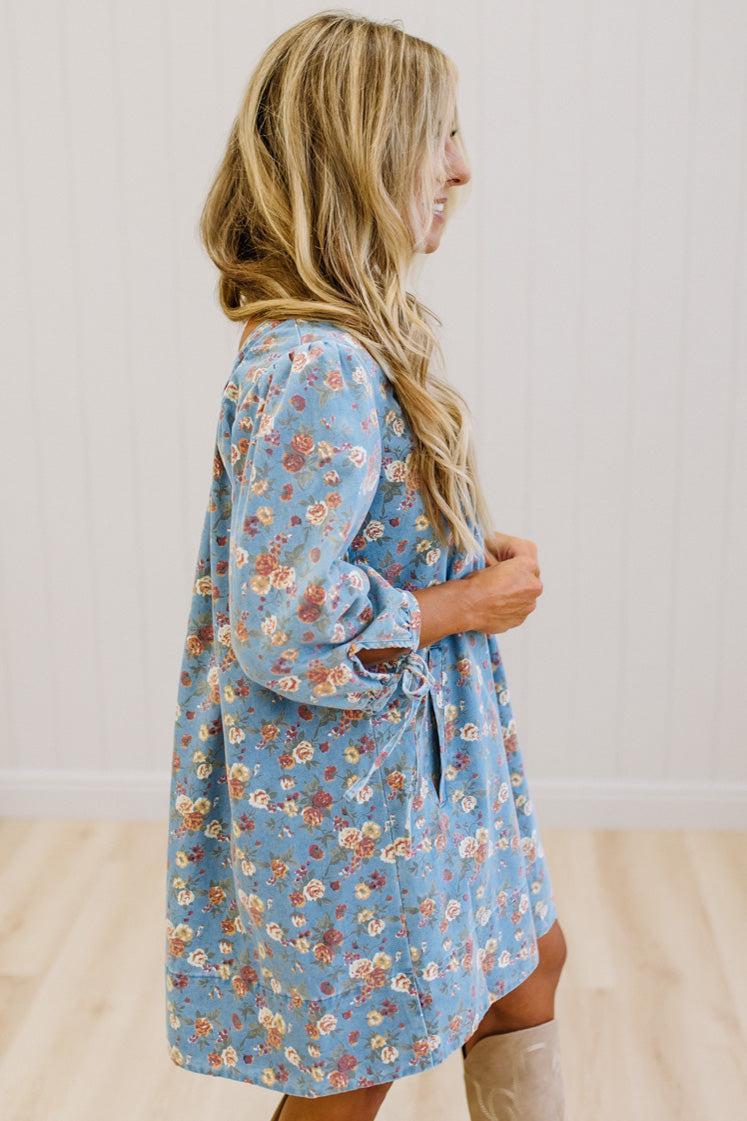A woman wearing a light blue floral robe with three-quarter sleeves and a drawstring waist, standing in a room with light wood floors and white paneled walls.
