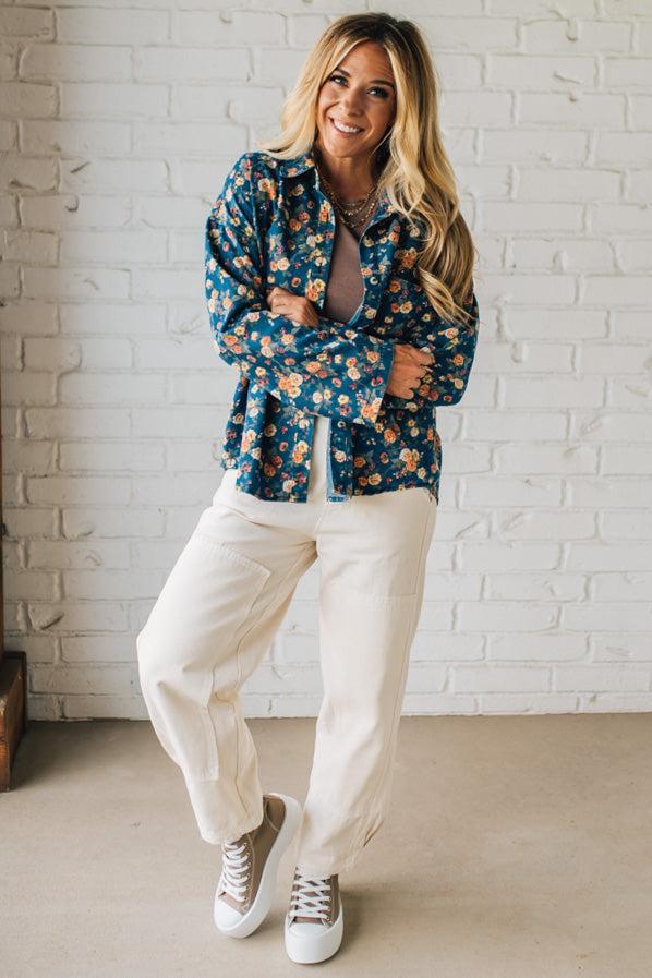 Woman wearing a floral blouse and white pants against a white brick wall.