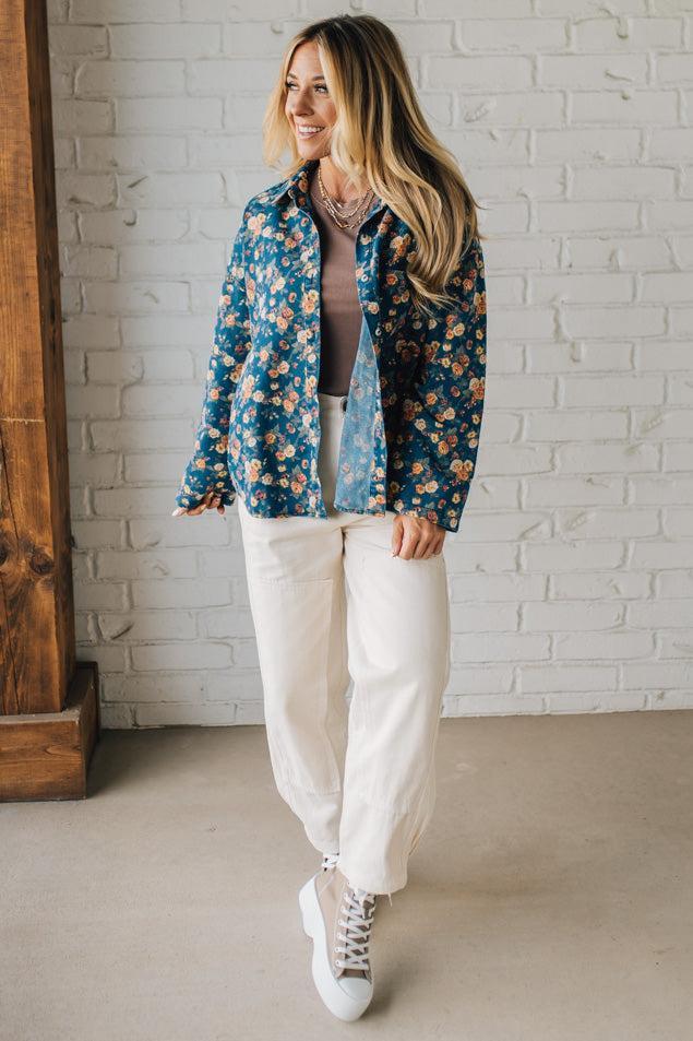 Woman wearing a floral jacket, gray top, white pants, and white sneakers against a white brick wall.
