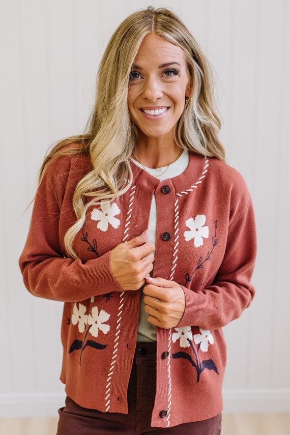 A smiling blonde woman wearing a rust-colored knit cardigan with white floral embroidery and cream stitched trim, layered over a white top and dark pants.