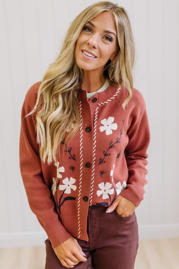 A woman modeling a rust-colored cardigan with white floral embroidery and a white top, paired with dark brown wide-leg pants and white pointed-toe heels, standing in a bright room with light wood floors and white paneled walls.