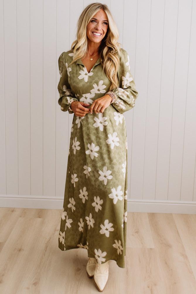 A woman modeling a long-sleeve olive green maxi dress with a white floral pattern, standing in front of a white paneled wall and light wood floor.