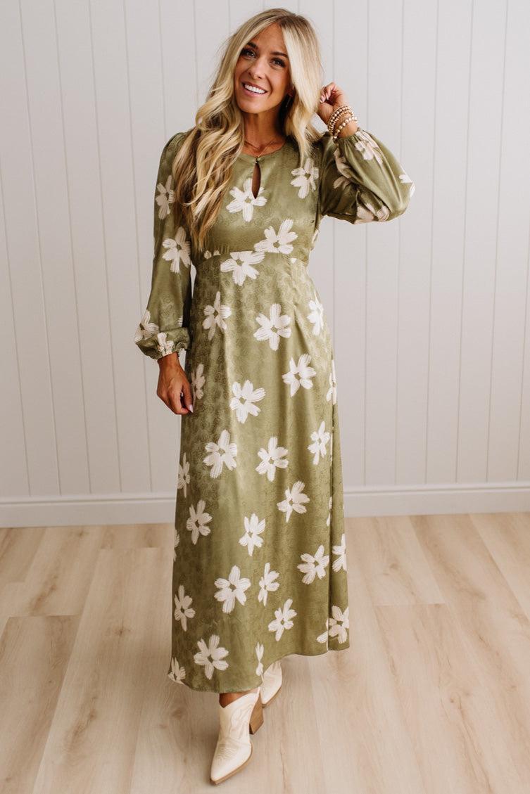 A woman modeling a long-sleeve olive green maxi dress with a white floral pattern, standing in front of a white paneled wall and light wood floor.