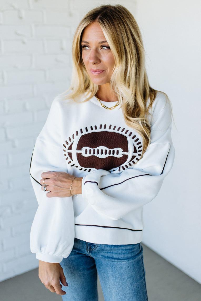 Woman wearing a white sweatshirt with a graphic design, standing against a white brick wall.