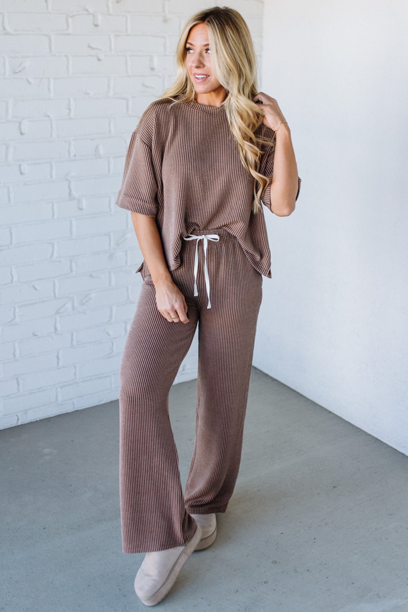 Woman wearing a brown ribbed outfit with wide-leg pants against a white brick wall.