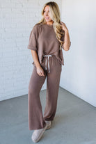 Woman wearing a brown ribbed outfit with wide-leg pants against a white brick wall.