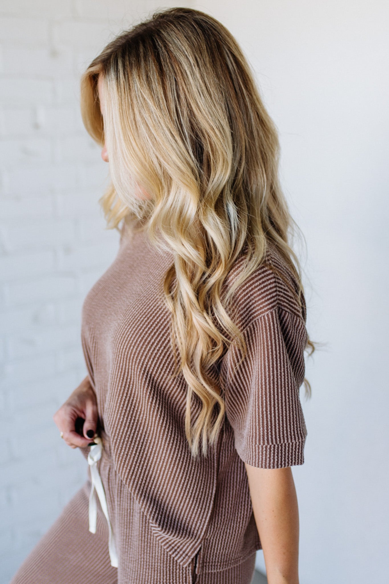 Woman with long, wavy blonde hair wearing a brown ribbed sweater against a white background