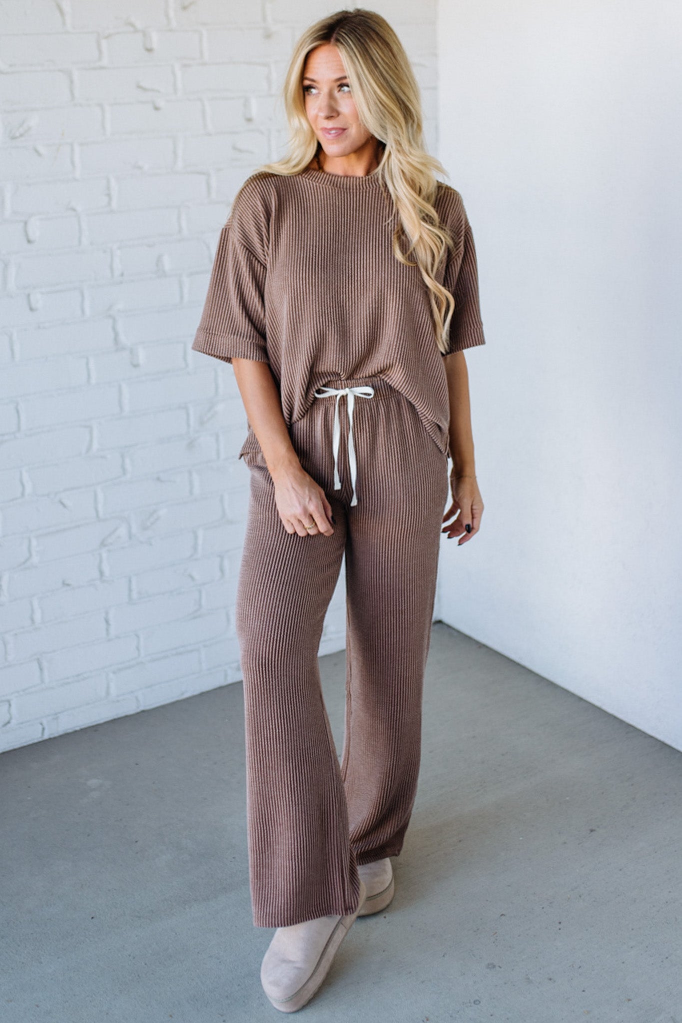 Woman wearing a brown outfit standing against a white brick wall.
