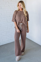 Woman wearing a brown outfit standing against a white brick wall.