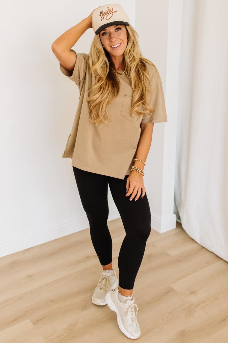 Woman wearing a beige t-shirt, black leggings, and white sneakers with a white cap indoors.