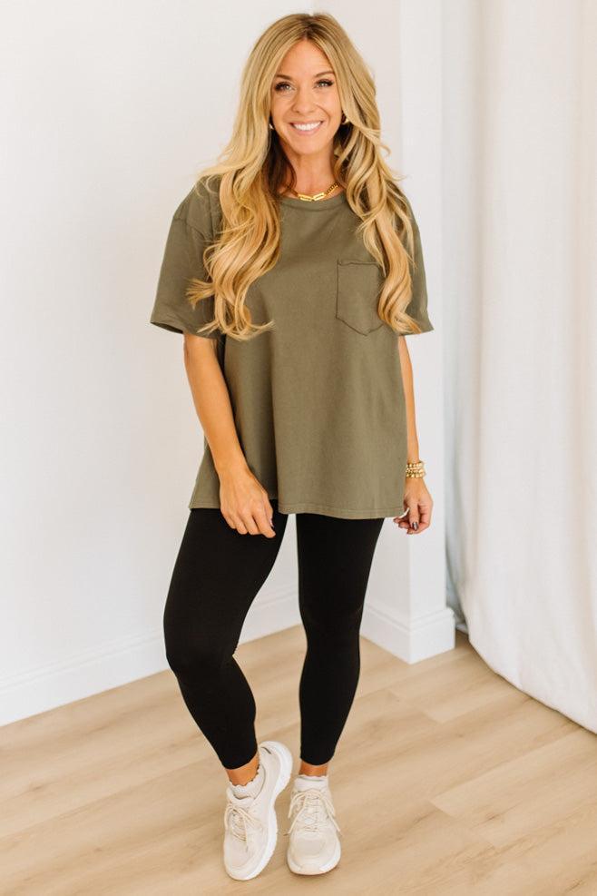 Woman wearing an olive green t-shirt, black leggings, and white sneakers in a minimalistic setting.