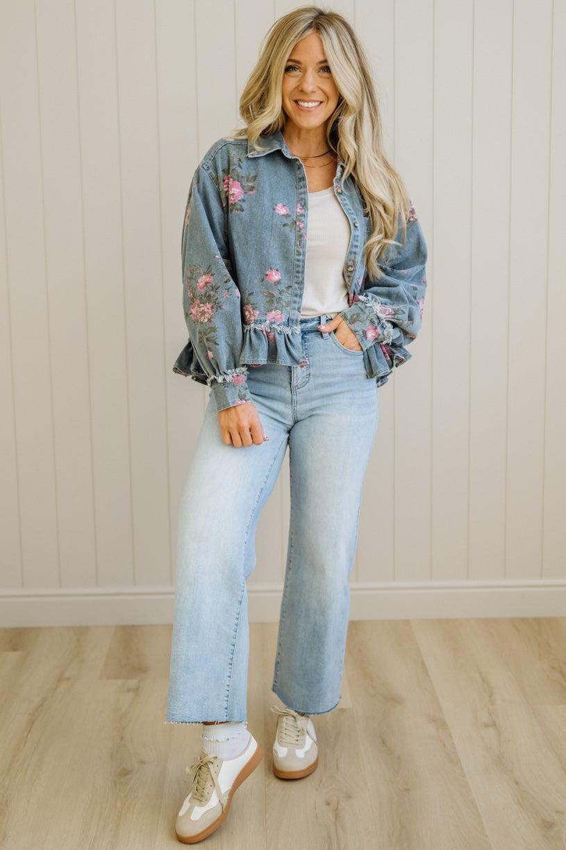 Woman wearing a floral denim jacket, white top, and light blue jeans indoors.