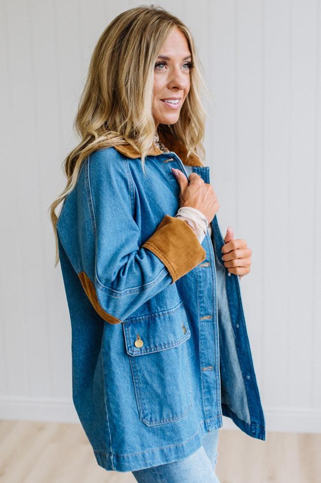 A woman wearing a blue denim jacket with brown suede-like trim on the collar and cuffs, standing in a bright indoor setting with a white paneled wall.