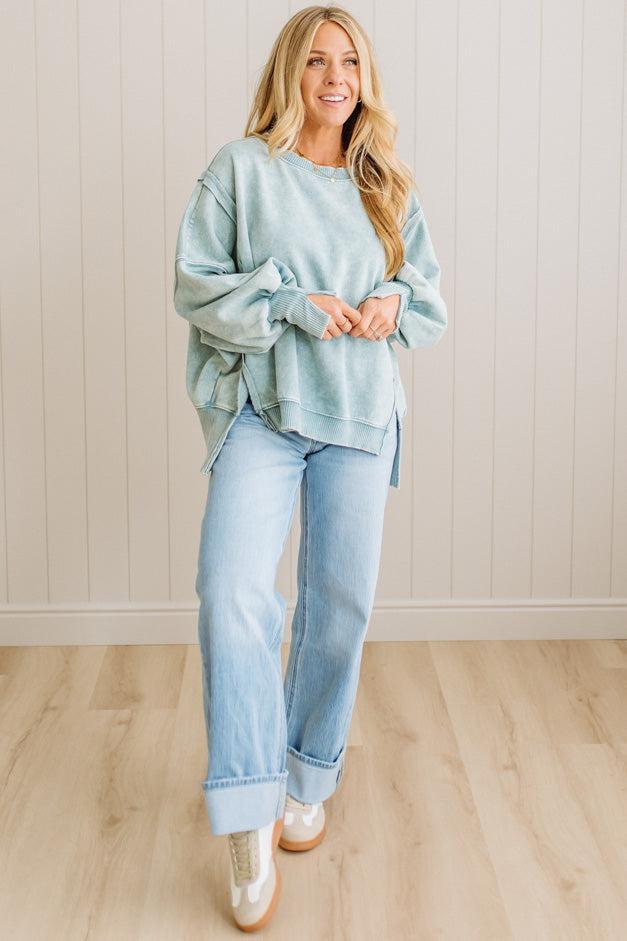 Woman wearing a light blue sweater and jeans standing in a room with wooden flooring and a white wall.