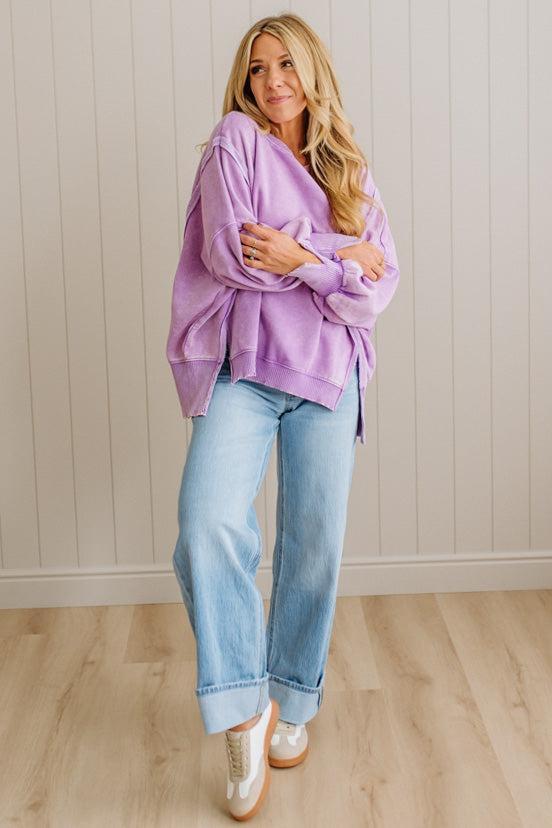 Woman wearing a purple sweater and blue jeans standing against a light wooden paneled wall.