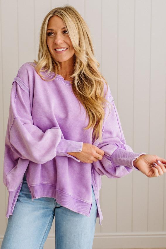 Woman wearing a purple sweater and blue jeans against a white paneled wall.