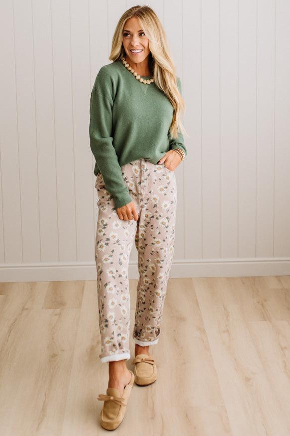 A woman stands in a room with light wood floors and white paneled walls, wearing a sage green long-sleeve knit sweater, cream pants with a small floral print, and tan slip-on shoes.