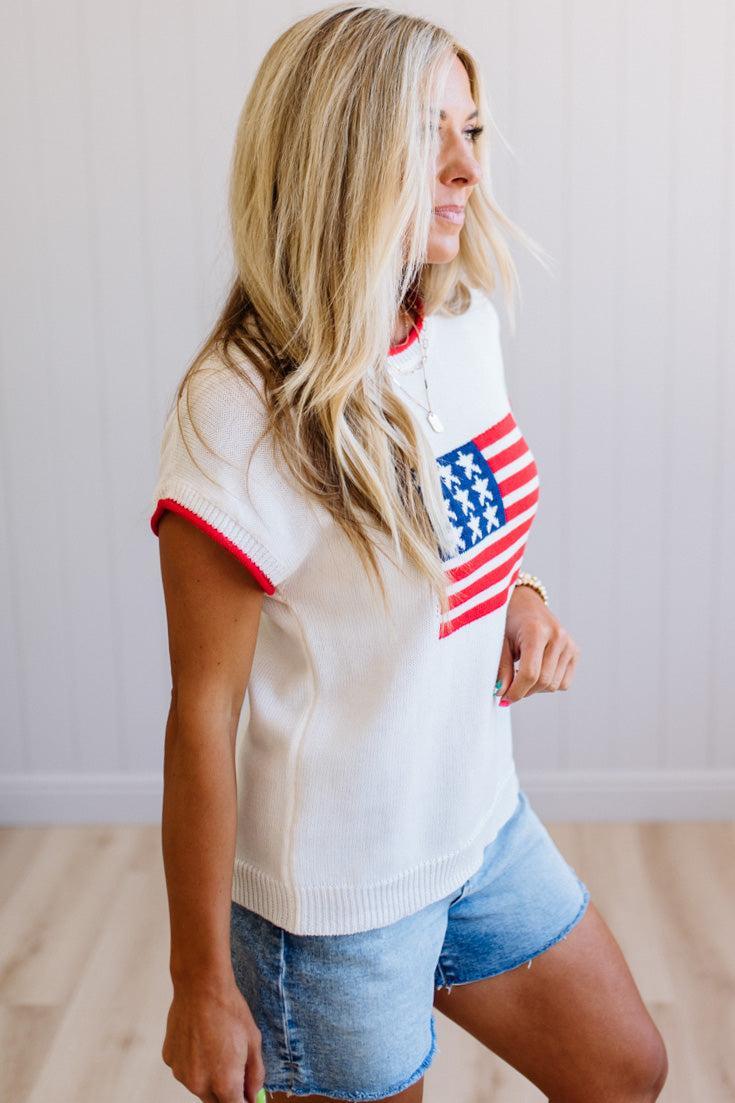 Woman wearing a white sweater with an American flag design and denim shorts indoors.
