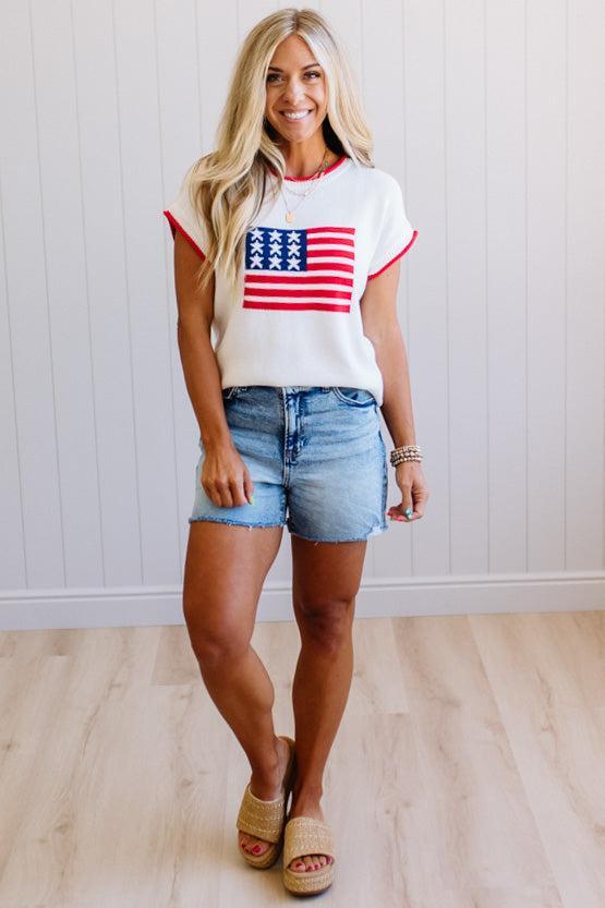 Woman wearing a white t-shirt with an American flag design and denim shorts indoors.