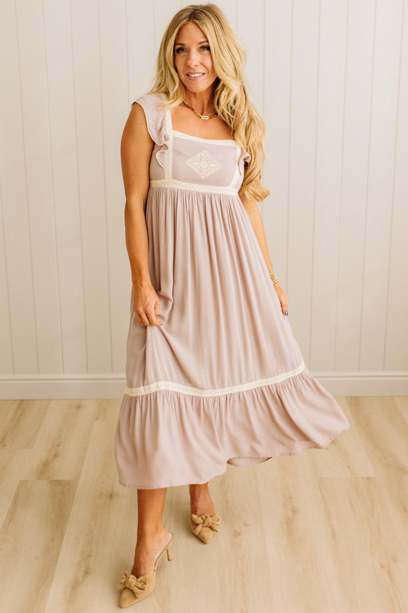 Woman wearing a light pink dress with white lace details in a room with wooden flooring and a white wall.