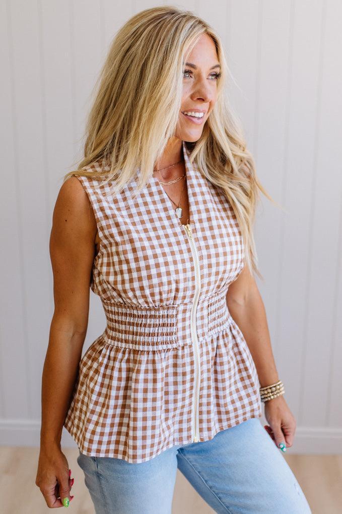Blonde woman wearing a mocha and white gingham print sleeveless top with smocked waistline and centered gold zipper front.