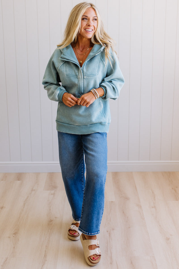 Woman wearing a light blue jacket and jeans standing against a white paneled wall.