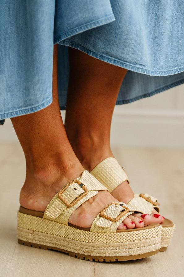 Beige sandals with espadrille sole worn with a blue denim skirt on a light wooden floor.