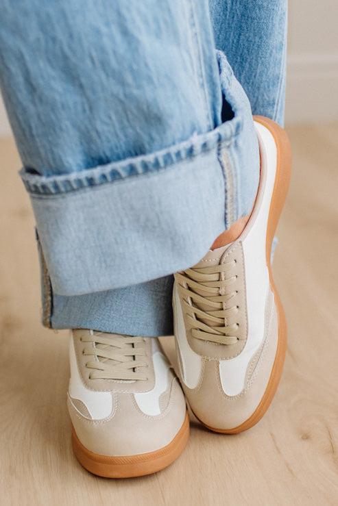 Beige sneakers with white accents and brown soles worn with light blue jeans on a wooden floor.