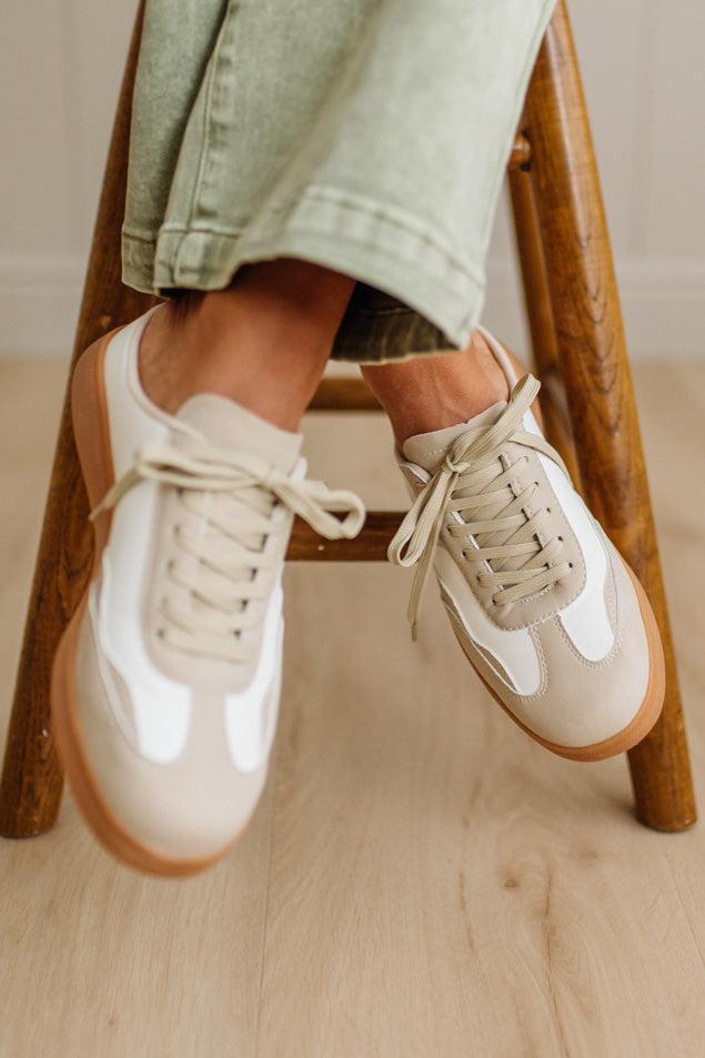 White and beige sneakers worn by a person sitting on a wooden stool.