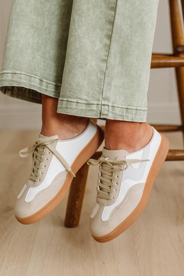 White sneakers with brown soles worn by a person sitting on a wooden stool.