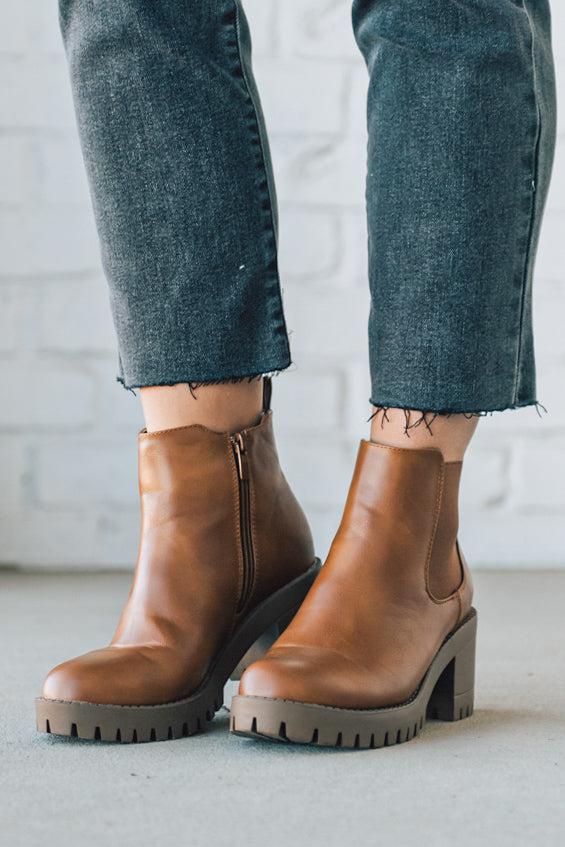 BROWN LEATHER BOOTS WITH BLACK CROP JEANS AGAINST A WHITE BRICK WALL.