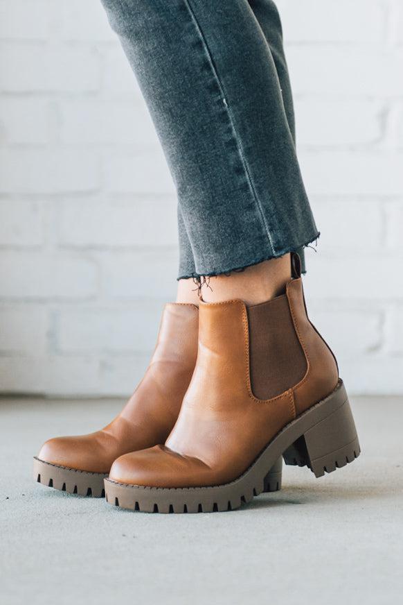 BROWN LEATHER BOOTS WITH BLACK CROP JEANS AGAINST A WHITE BRICK WALL.