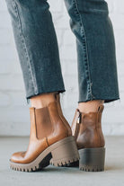 BROWN LEATHER BOOTS WITH BLACK CROP JEANS AGAINST A WHITE BRICK WALL.