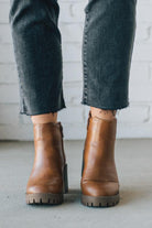 BROWN LEATHER BOOTS WITH BLACK CROP JEANS AGAINST A WHITE BRICK WALL.