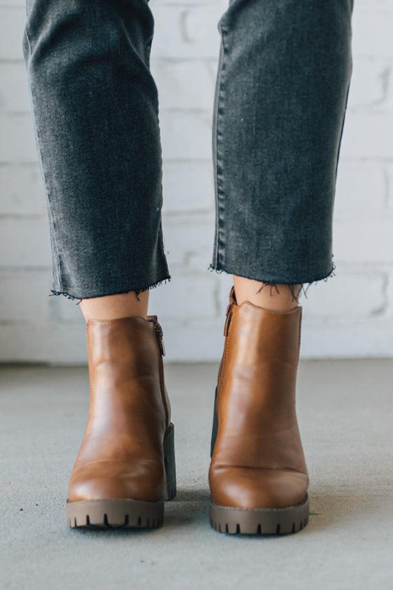 BROWN LEATHER BOOTS WITH BLACK CROP JEANS AGAINST A WHITE BRICK WALL.